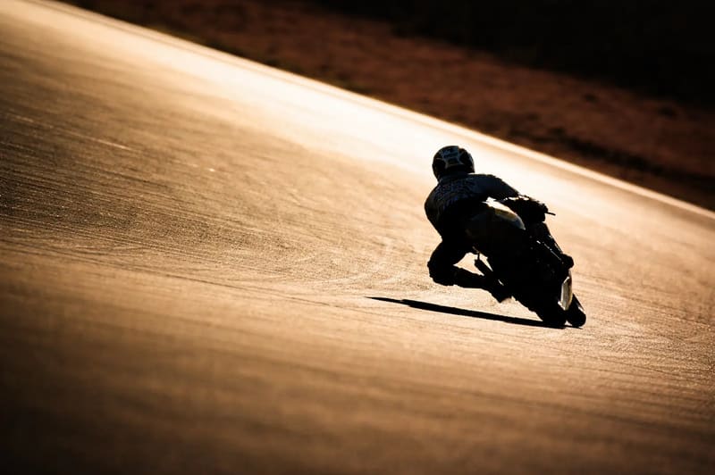 A motorcycle racer leaning low through a sweeping bend on a sunlit track at sunset